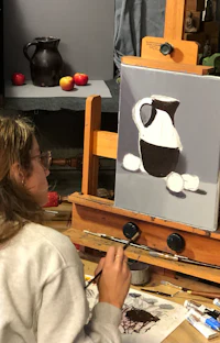 Beginner artist painting a still life of a brown jug and red apples. 